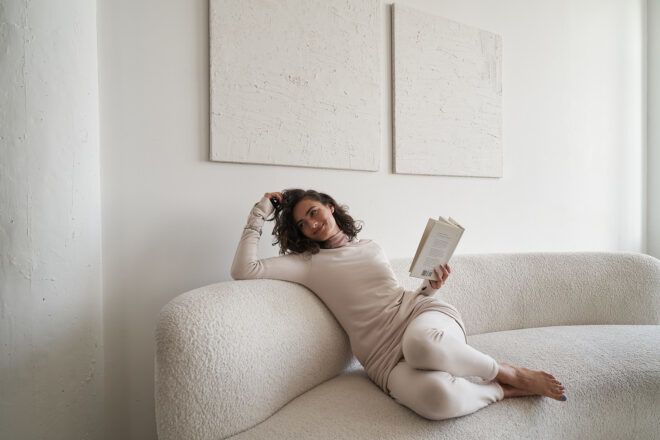 A woman wearing a pink long-sleeve shirt and white legging lounges on a white sofa with one hand in her hair and the other hand holding a book