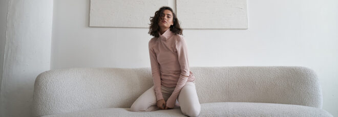 A woman waring a pink long sleeve shirt with white pants sits on her knees on a white sofa