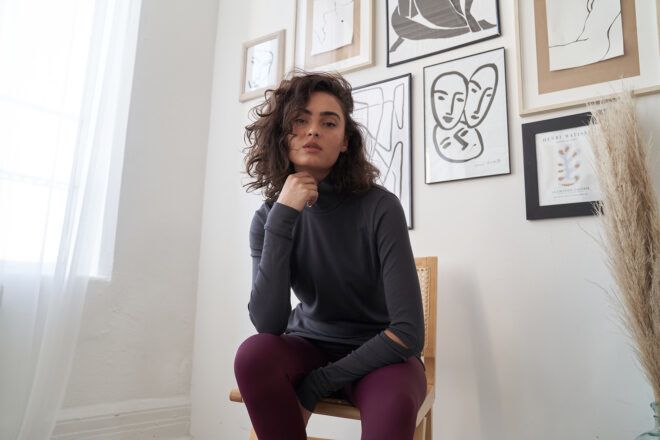 A woman wearing a grey long-sleeved shirt and maroon pants sits on a wooden chair with her hand underneath her chin and the other underneath her knee