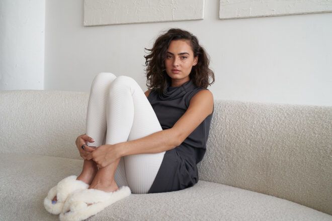 A woman wears a gray sleeveless shirt with white leggings and fluffy scandals with her knees tucked to her chest on a sofa