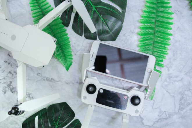 A white phone next to a drone remote and a white drone with fake leaves surrounding it