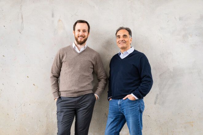 Two men stand against a gray wall
