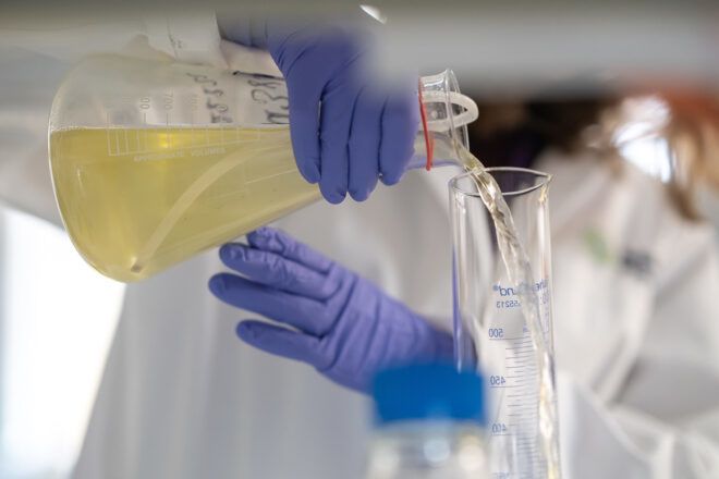 A person holds a beaker with a light yellow fluid to pour it into a clear beaker