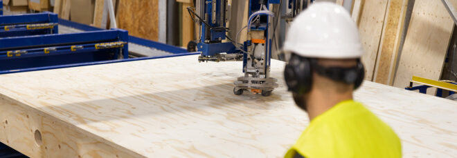 A wooden plank being cut with a machine and supervised by a man wearing a white hat and yellow hazard jacket