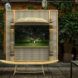 Installation in a courtyard with a sleek bamboo bench in front of a large earthy toned frame with intricate patterns and detailing. The frame encompasses a screen that shows two people in a grassy field surrounded by monkeys