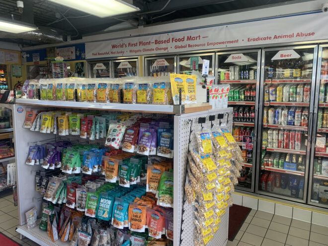 The inside of a convenience market with a wall of drinks in refrigerators and stands of chips and snacks