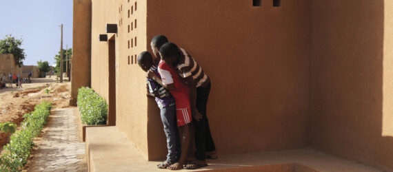 Three children peeking out from the corner of a red brick masonry building