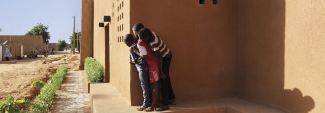 Three children peeking out from the corner of a red brick masonry building
