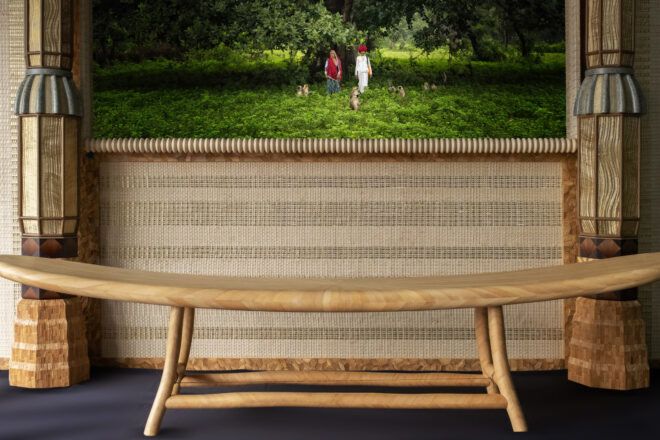 Sleek bamboo bench in front of a large earthy toned frame with intricate patterns and detailing. The frame encompasses a screen that shows two people in a grassy field surrounded by monkeys