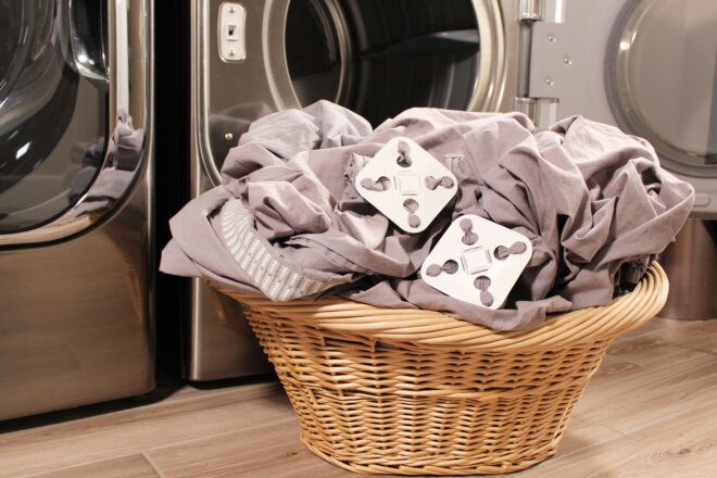 A pile of sheets with two white squares attached to it sits in a laundry basket beside a laundry machine