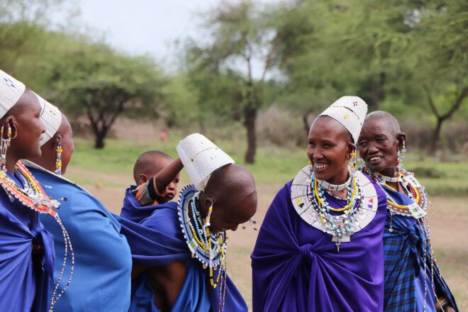 A group of Kenyan women wearing purple and blue robes with white caps and smiling at each other