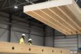 Two people wearing white helmets and yellow hazard suits watching a wood part being lifted in the air