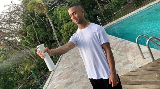 An 18-year-old man wearing a white shirt with black bottoms standing beside a pool and spraying from a white canister