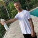 An 18-year-old man wearing a white shirt with black bottoms standing beside a pool and spraying from a white canister