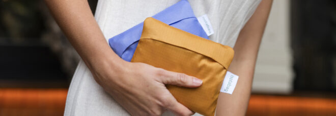 A hand holding two tote folded down into rectangles, one purple and one orange