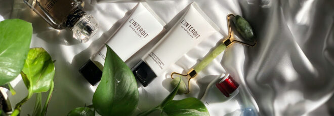 A flat lay on a white cloth of leaves, white cream bottles and a green face roller