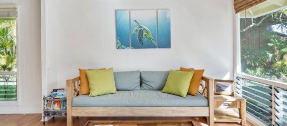A sofa with blue, yellow and orange cushions set beside an open window and set below a painting of a turtle