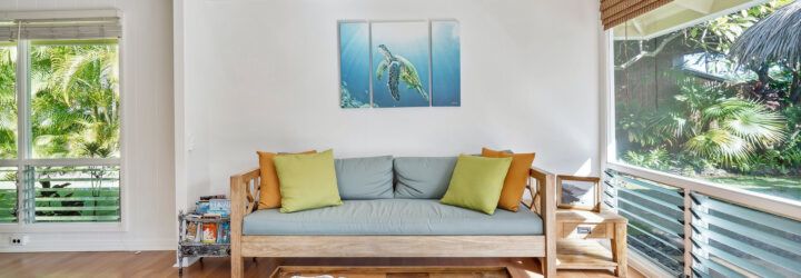 A sofa with blue, yellow and orange cushions set beside an open window and set below a painting of a turtle