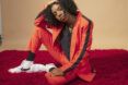 A woman with Afro hair wearing an orange suit with white sneakers with one leg propped up