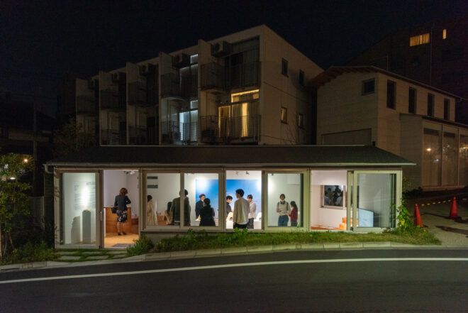 A building illuminated with light from within of people viewing an exhibit on the ground floor