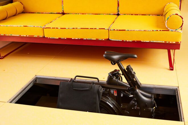 A black exercise bike hidden in a space underneath the flooring with a yellow sofa on the ground floor