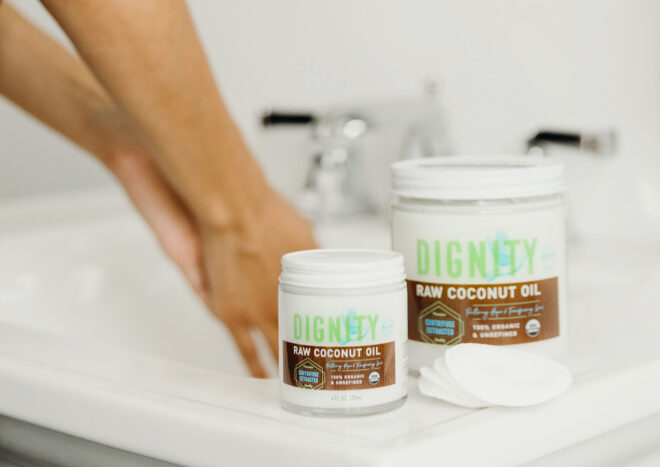 Two jars of Dignity raw coconut oil, in the background are two hands washing under a sink