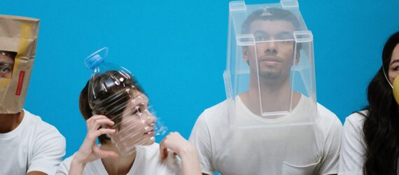 Four people in a row, the first wearing a brown paper bag over his head, the next with a plastic bottle over her head, the next a plastic bin over his head and the last wearing a yellow face mask