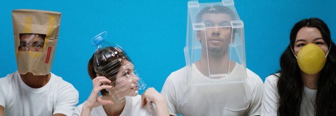 Four people in a row, the first wearing a brown paper bag over his head, the next with a plastic bottle over her head, the next a plastic bin over his head and the last wearing a yellow face mask