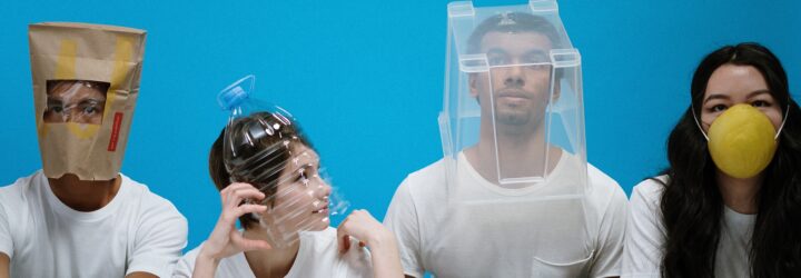 Four people in a row, the first wearing a brown paper bag over his head, the next with a plastic bottle over her head, the next a plastic bin over his head and the last wearing a yellow face mask