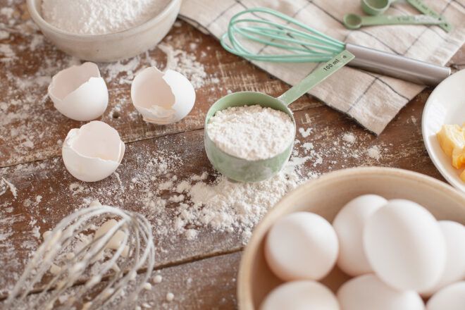 A measuring spoon with flour, two whisks, eggs and empty empty egg shells on a wooden counter