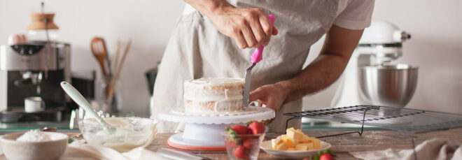A hand icing a cake on a messy counter with baking utensils