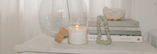 A white lit candle set on a table stand in front of a stack of books and a glass jar