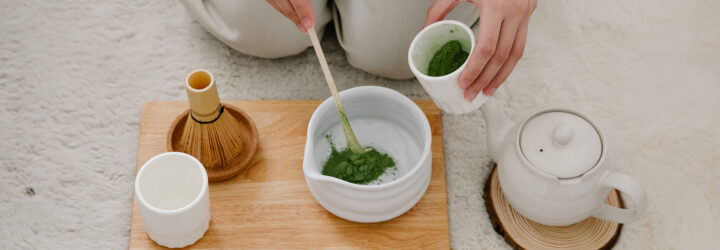 A person kneeling and preparing matcha in a cup and a bowl with a teapot and mixer beside them