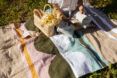 An up-close shot of a person sitting reading a book with a woven basket beside them, sitting on a blanket on grass