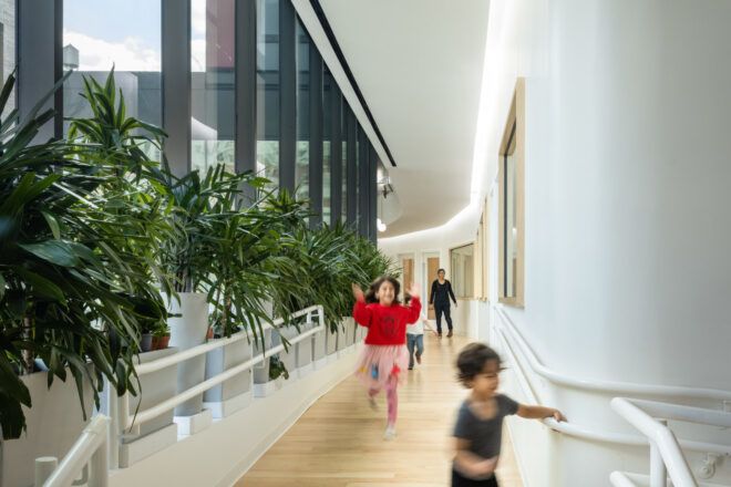 Children running through a narrow corridor with tall windows flanked by large plants