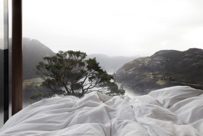 A white blanket of a bed and a window that overlooks a mountain range