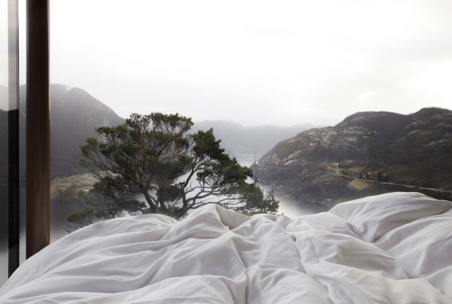 A white blanket of a bed and a window that overlooks a mountain range