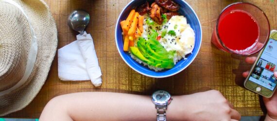 An arm of a person and the other arm holding a phone with a bowl of food, a red punch drink and a hat beside them