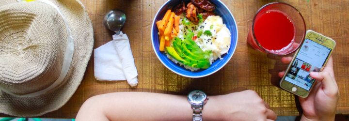 An arm of a person and the other arm holding a phone with a bowl of food, a red punch drink and a hat beside them