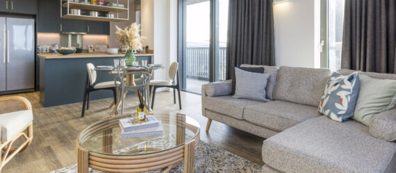 Modern apartment interior featuring a seating and dining area with neutral colored furniture and a kitchen with dark green cabinetry