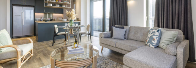 Modern apartment interior featuring a seating and dining area with neutral colored furniture and a kitchen with dark green cabinetry