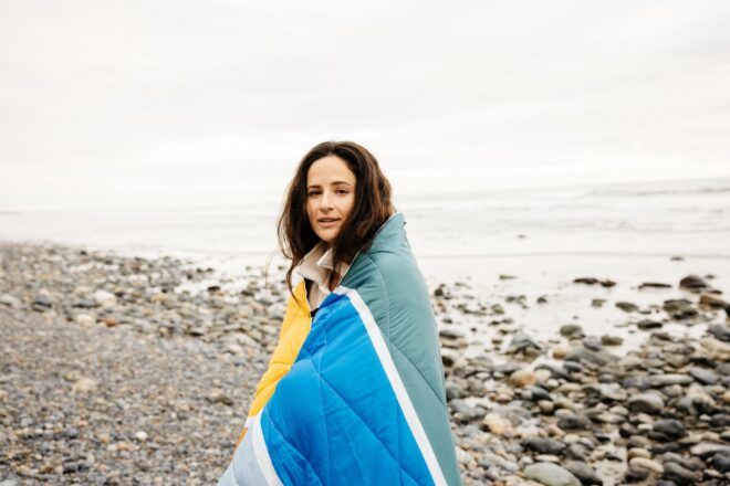 A woman wrapped in a puffer blanket standing beside an ocean