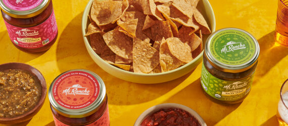 A basket of tortilla chips, jars of salsa and a bowl of green salsa