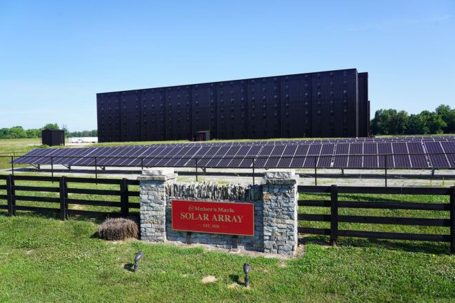 A stone sign that reads the Maker's Mark Solar Array with a fence, solar panel, and black building behind it
