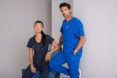 A man and a woman dressed in scrubs leaning against a gray wall