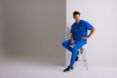 A man in blue scrubs sitting on a stool beside a white wall