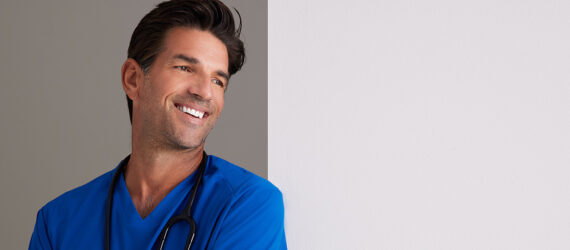 The top half of a man wearing a blue scrub leaning against a white wall