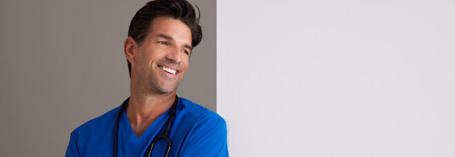 The top half of a man wearing a blue scrub leaning against a white wall