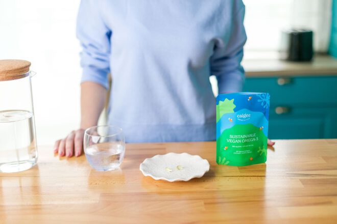 A person wearing a light blue sweater standing behind a wooden counter with a cup of water, white plate and Calgee Omega-3 package