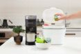 A hand putting a coffee pod into a compost bin with a coffee maker beside with a bowl of coffee pods